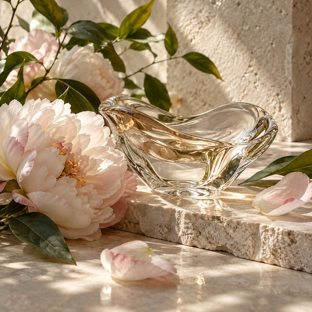 Editorial artwork for Ane Ayo: soft peony petals and green leaves around a clear sculptural glass on pale limestone.